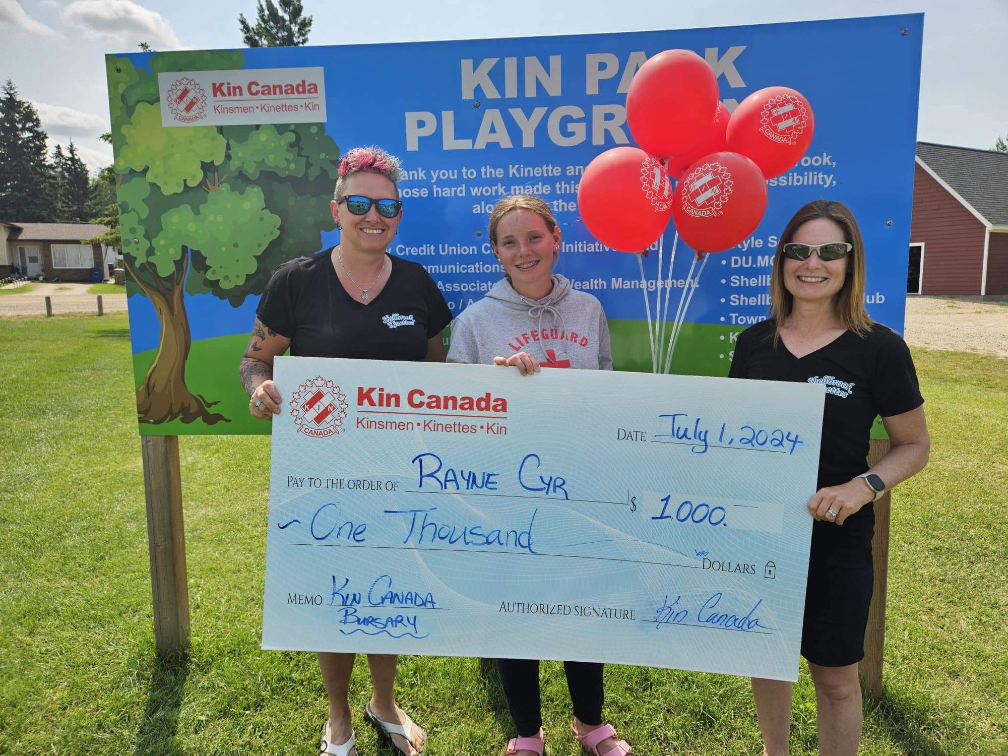 Three people holding a large cheque looking at the camera smiling with balloons in front of the Kin Park Playground sign